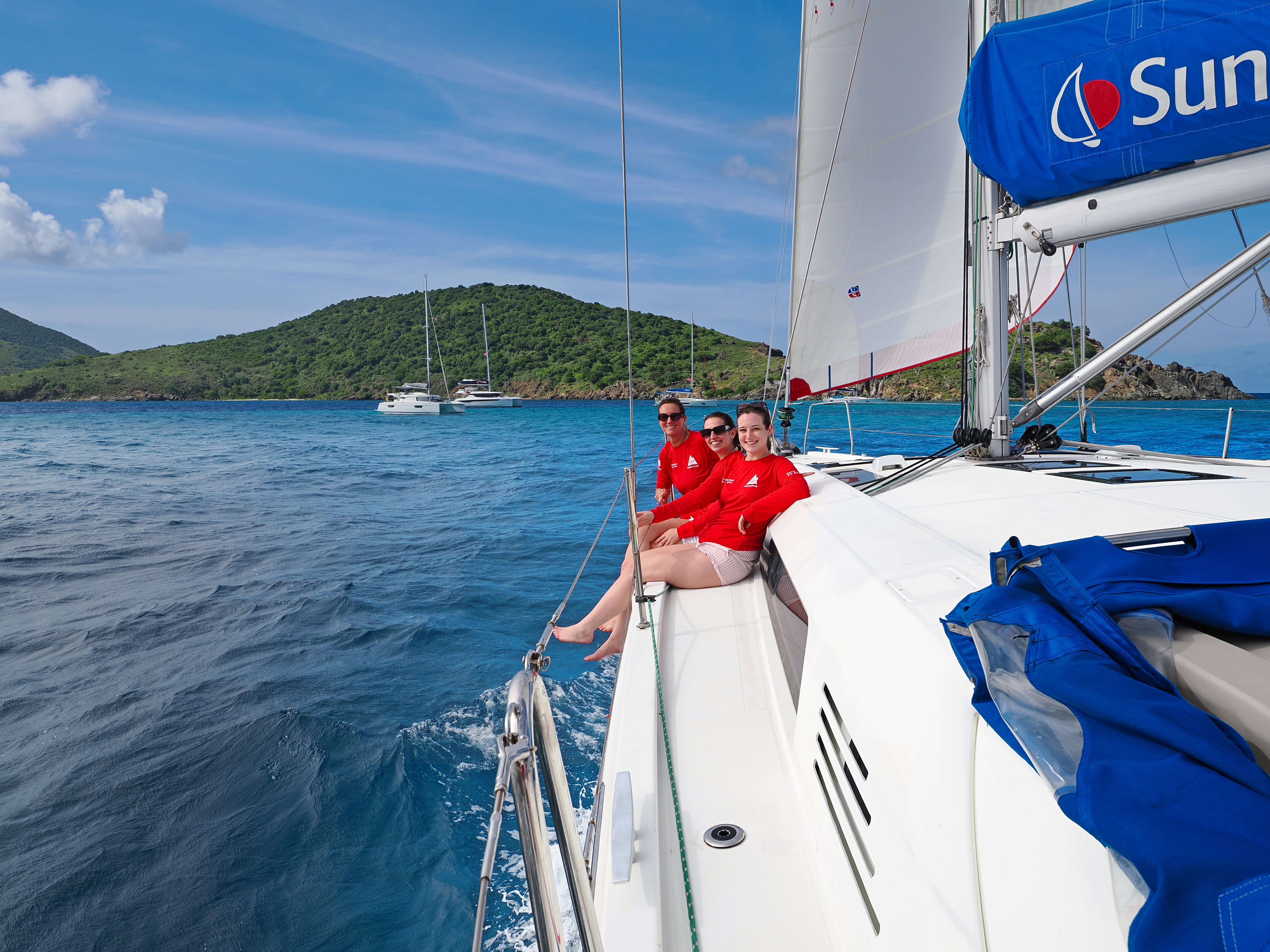 Three people in red tops sit on a sailing catamaran near Sandy Spit, British Virgin Islands, with turquoise water and green hills behind them.